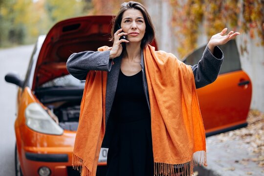 Woman calling for roadside assistance beside an orange car with its hood open, gesturing in frustration after a breakdown on an autumn street