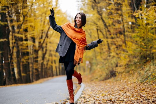 Woman in an orange scarf balancing with outstretched arms on a leaf-strewn park path beneath golden autumn trees