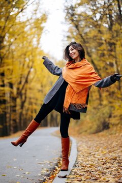 Woman in an orange scarf balancing with arms outstretched on a leaf-strewn forest path, enjoying a bright autumn day