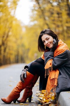 Woman in a gray coat and orange scarf, sitting beside an autumn path and enjoying a quiet moment among golden trees