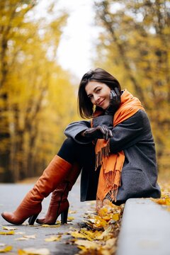 Woman sitting beside a leaf-covered path, smiling in a gray coat, orange scarf and brown leather boots beneath golden autumn trees