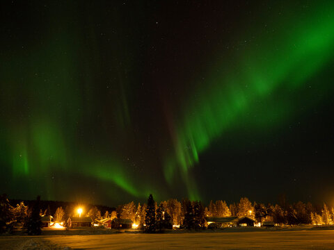 Aurora borealis northern lights glowing in the night sky over a snow-covered forest and illuminated winter village in Lapland