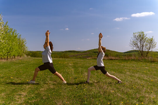 Two women performing yoga lunges with arms raised in sunny green meadow surrounded by rolling hills and scattered trees, casual workout clothing, spring daylight