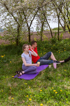 Young woman and man sitting on purple yoga mat under blossoming trees in grassy meadow enjoying relaxed outdoor break with reusable bottles and takeaway cup while smiling and chatting