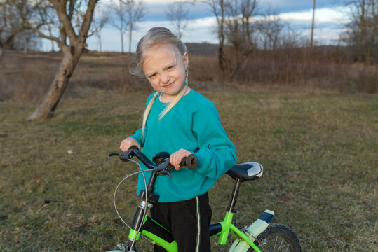 Young blonde girl expressing a disgruntled or playful emotion while standing with her small green bicycle in a rural outdoor setting during autumn, looking directly at the viewer