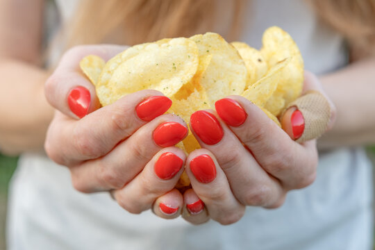 Woman's manicured hands with bright red nails cupping a large portion of crispy, seasoned potato chips, close up conveying casual indulgence, craving and savory crunch