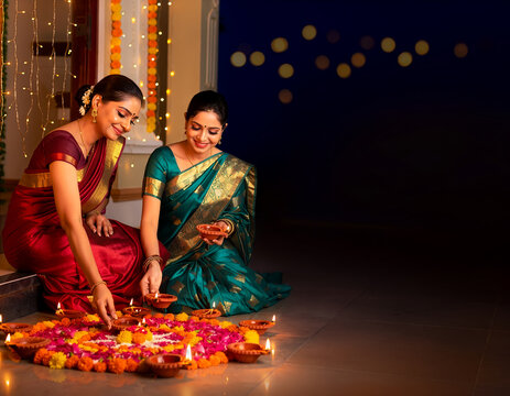 Indian women light diyas around a floral rangoli for Diwali