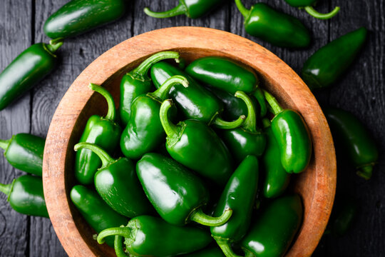 Jalapeno peppers in wooden bowl with additional pieces around on dark surface. Fresh green chili natural food layout