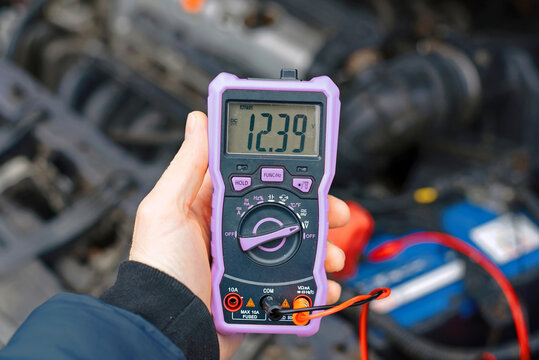 Drivers hand holding multimeter reading 12.39 volts connected to car battery in cold winter season. Voltage tester showing 12.39 volts - partially charged, but still works. Winter car battery testing