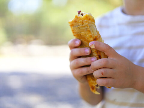 Child holding a bitten flaky pastry in both hands outdoors during a snack break, eating a bakery treat that brings convenient baked goods into children's diets as a handheld portion between meals.