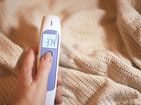 Digital thermometer reading 37.4&deg;C is held against a knitted blanket during a body temperature check, a common home healthcare task when monitoring possible fever symptoms and caring for illness.