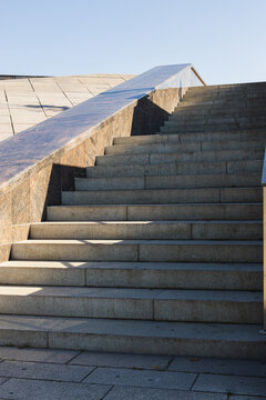 Wide concrete staircase with angular stone walls and metal railing under a clear blue sky in an outdoor modern architectural setting