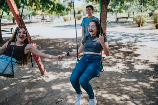 Young adult friends swing, laugh and hold hands together at an outdoor park playground, celebrating youth, leisure and carefree fun.