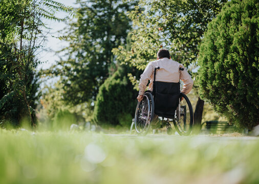 Man uses a wheelchair independently along a sunny park path, representing mobility, disability inclusion, accessibility and active outdoor living.