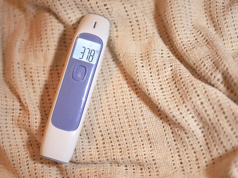 Digital thermometer showing 37.8&deg;C rests on a knitted blanket, with its backlit screen displaying the elevated body temperature reading used in home healthcare for fever monitoring during illness.