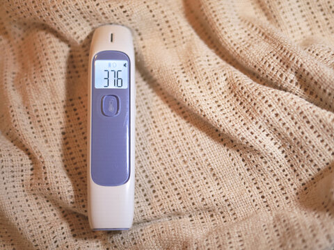 Digital thermometer reading 37.6 rests on a knitted blanket, its illuminated screen showing a body temperature measurement for home healthcare and fever monitoring when checking symptoms of illness.