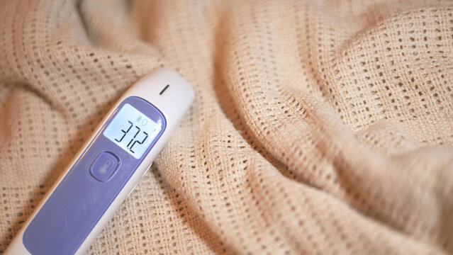 Digital thermometer reading 37.2 rests on a knitted blanket, a handheld medical device for home healthcare and body temperature checks with an illuminated display for monitoring possible fever.