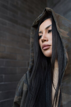 Close-up portrait of a beautiful Asian woman with long straight dark hair wearing a camouflage hoodie outdoors near a dark brick wall background. Modern urban street fashion look.