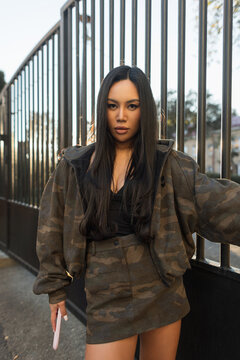 Confident Asian woman in trendy camouflage jacket and mini skirt looking at camera while posing near metal gate in warm evening light. Portrait of stylish brunette girl holding smartphone