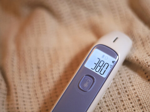 Digital thermometer displaying 38.0&deg;C rests on knitted fabric, a home healthcare device for monitoring fever and checking elevated body temperature during illness, with a backlit screen in focus.
