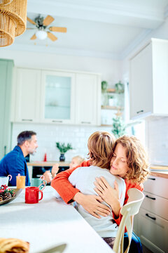 Mother embracing her son at the kitchen table, sharing a tender family moment as father and child talk in the background