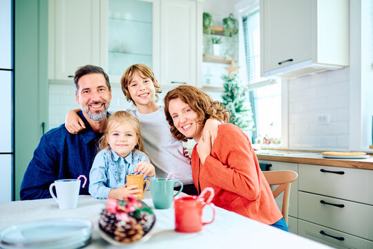 Smiling parents and children gathering around the kitchen table, sharing warm drinks beside a decorated Christmas tree