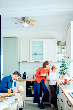 Family members sharing a festive morning in the kitchen, with a mother assisting her son at the counter and a father cradling their baby