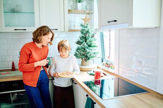 Mother and young son sharing a holiday baking moment, holding a homemade pie beside a decorated Christmas tree in their bright kitchen