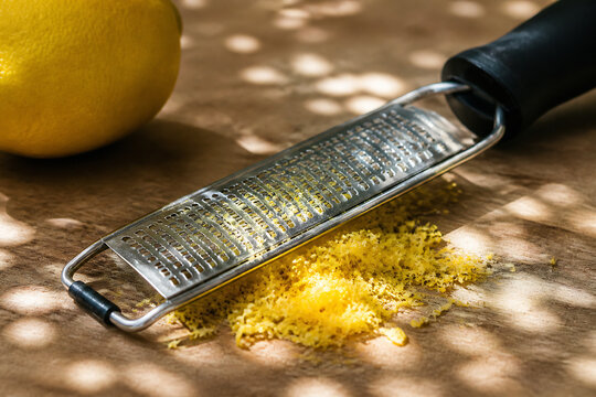 A lemon zester with grated yellow zest on a wooden surface under dappled sunlight.