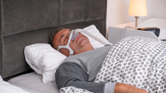 Middle-aged man sleeping in bed wearing a CPAP mask, sleep apnea device to treat obstructive sleep apnea and prevent breathing pauses during the night.