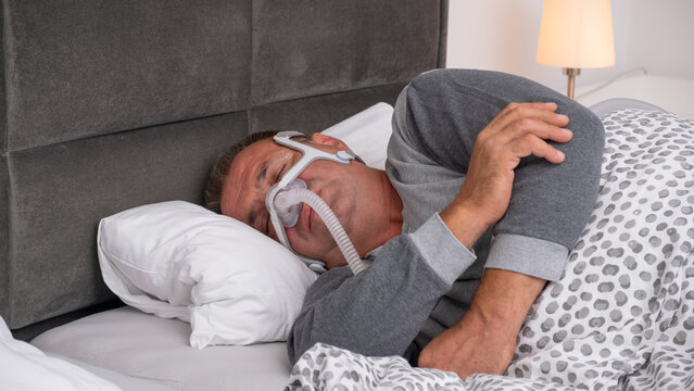 Middle-aged man sleeping in bed wearing a CPAP mask, sleep apnea device to treat obstructive sleep apnea and prevent breathing pauses during the night.
