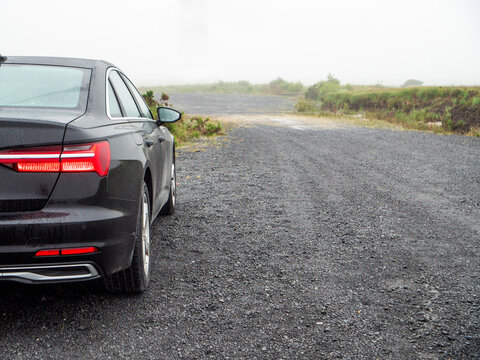 Black sedan parked on gravel road near green grass. Sky is overcast and gray. Car shows modern design with sleek lines. Image suitable for travel, automotive, or roadside themes
