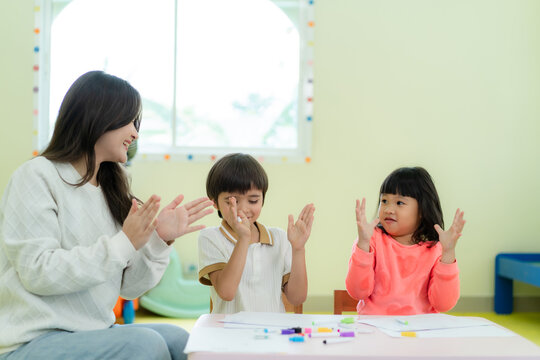 A happy Asian female teacher in a white sweater claps hands with young children during an interactive fun activity in a kindergarten classroom.