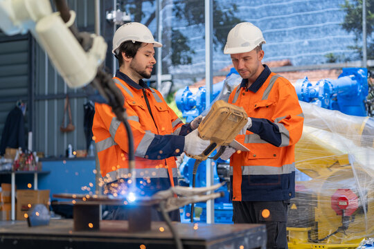 Two Caucasian male engineers in orange safety uniforms and hardhats control an automatic welding robot arm using a device inside a factory workshop.