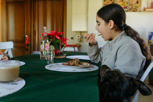 Young girl eating dessert at dining table while dog watches longingly for food