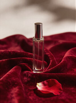 Clear square perfume bottle with silver cap on draped burgundy velvet with a single rose petal