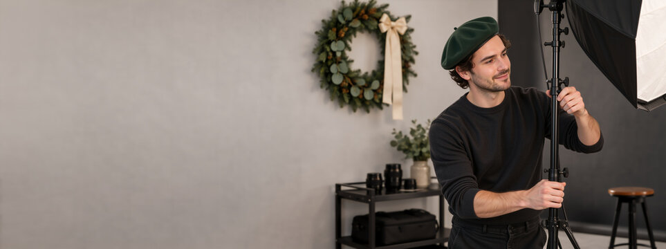 Professional photographer adjusting softbox lighting in studio with holiday wreath. Young man setting up photoshoot. Copy space for text