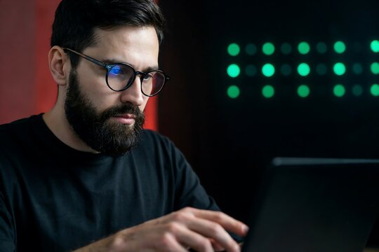 Bearded man in glasses working on laptop in dark office with glowing green server lights in background