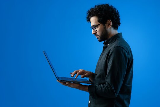 Young man with curly hair using laptop computer while standing against a solid blue studio background