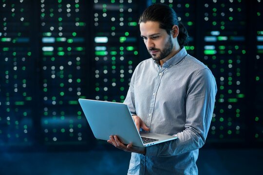 Professional technician using laptop while monitoring server racks in a modern dark data center facility room