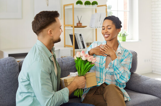 At home on sofa, in shares tender moment as man presents gift and flowers to girlfriend. Happy, smiling African American woman says a heartfelt thank you, showing gratitude and admiration