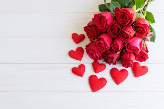 A generous bouquet of red roses sits beside scattered red hearts on a wooden surface.