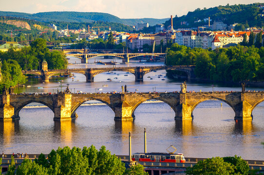 Golden hour aerial view from Letna Park over Charles Bridge, the Vltava River, and crowded historic Prague cityscape with arches, statues, boats, and warm evening light.