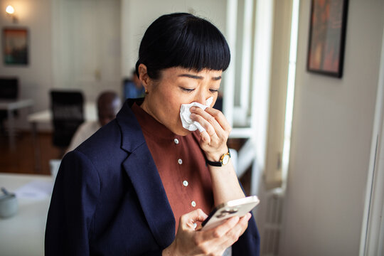 Sick businesswoman with tissue checking phone in office