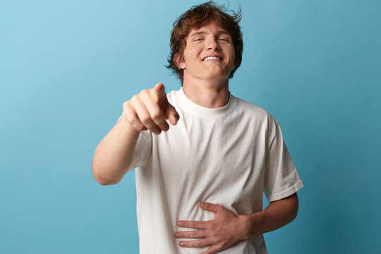 Smiling man in a casual white t shirt points at the camera, hand on stomach, mid-laugh, blue background, creating a playful, relatable vibe for marketing engagement and social campaigns.