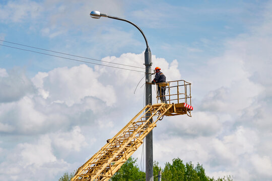 Worker in hard hat in aerial lift basket working on street light pole under blue sky. Utility technician on elevated platform performing maintenance on tall lamp post. Worker repairing street lighting