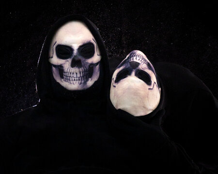 Two people wearing skull masks against a smokey black background.